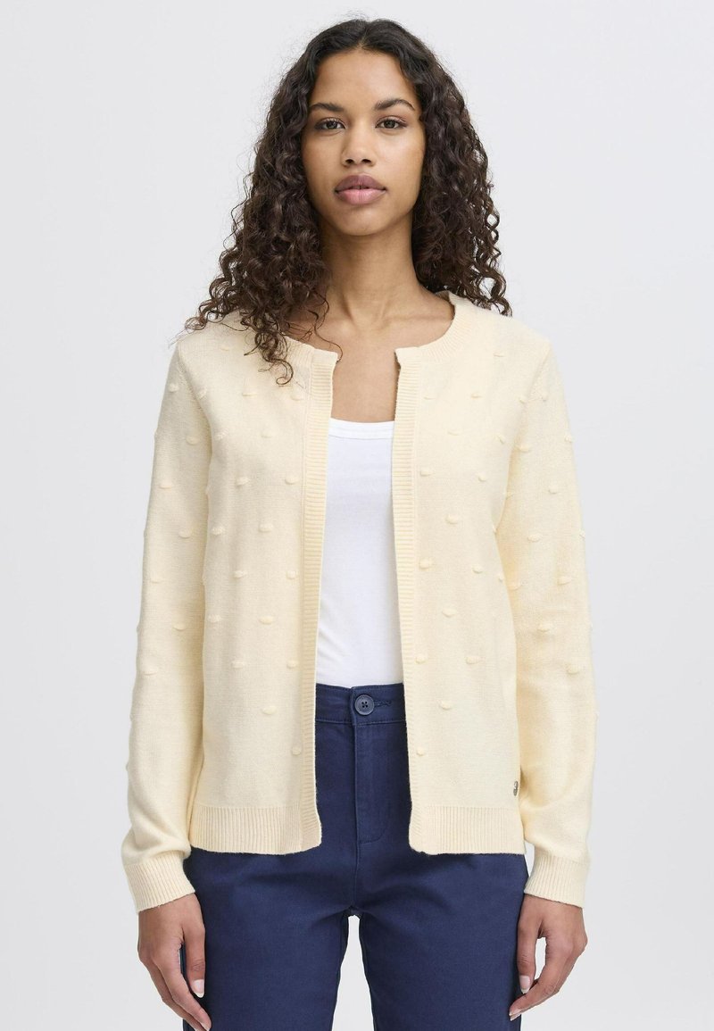 Světle žlutý cardigan s texturovanými bobble detaily, žebrovanými manžetami a lemem, nošený přes bílé tričko, v kombinaci s tmavě modrými kalhotami.