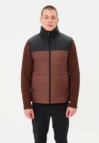 Braune gesteppte Weste mit schwarzen Schultern, hohem Kragen und Frontreißverschluss, kombiniert mit einem braunen, strukturierten Langarmshirt.