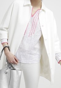 Weiße Oberbekleidung, die über einer bedruckten Bluse mit pinken Akzenten lagert, kombiniert mit schmalen weißen Hosen und einer silbernen, strukturierten Handtasche.