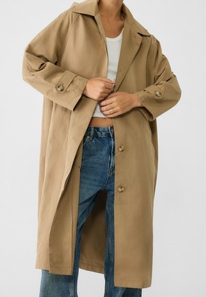Personne portant un long trench-coat beige par-dessus un débardeur blanc court et un jean bleu, ajustant les boutons avant du manteau.