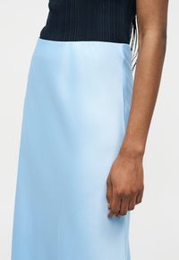 Γυναίκα που φοράει φούστα ανοιχτού μπλε με ψηλή μέση και μαύρο ριμπ αμάνικο τοπ, με το δεξί της χέρι και χέρι χαλαρό στο πλάι.