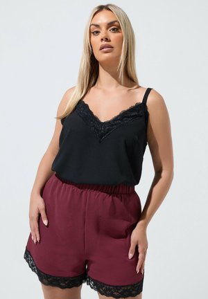 Kobieta ubrana w czarny top z koronkową lamówką i wysokim stanem bordo szorty z czarną koronkową lamówką, stojąca na jednolitym jasnym tle.