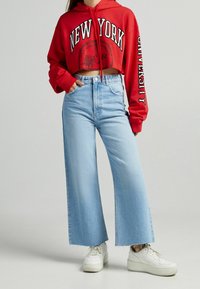 Sudadera corta roja con estampado de "NUEVA YORK", combinada con jeans de pierna ancha azul claro y zapatillas blancas, con detalles de dobladillo deshilachado.