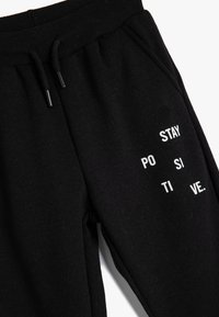 Schwarze Jogginghose aus weichem Stoff mit einem Kordelzugbund und bedrucktem "STAY POSITIVE"-Text in Weiß auf der Vorderseite.