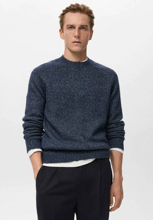 Maglione lavorato a maglia blu navy con scollatura a girocollo, finitura testurizzata e orlo a costine. Indossato sopra una camicia bianca a strati, abbinato a pantaloni scuri.