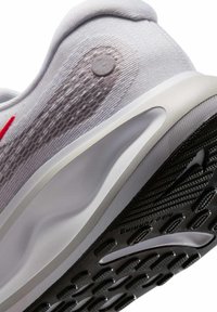 Sneaker grigio chiaro con tomaia in rete testurizzata, accento rosso e suola in gomma nera caratterizzata da motivi a scanalatura circolare e un sottopiede liscio.