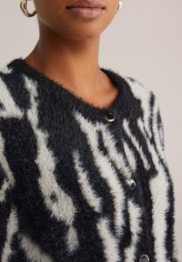 Svart och vit randig stickad cardigan med en fluffig textur, rund halsringning och metallknappar. Mönstret har en vågig, abstrakt design.