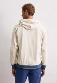 Młody mężczyzna stojący tyłem, ubrany w kremowego hoodie z ciemnoniebieskimi mankietami i pasem, w zestawieniu z niebieskimi spodniami na jasnym tle.