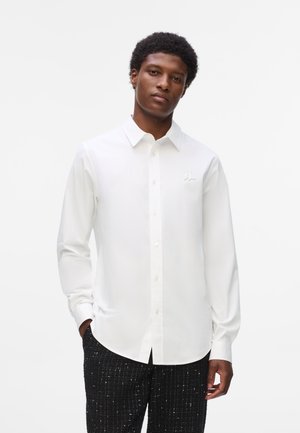 Chemise blanche à manches longues avec col, arborant un petit logo sur la poitrine, associée à un pantalon noir texturé. Fini en tissu lisse.