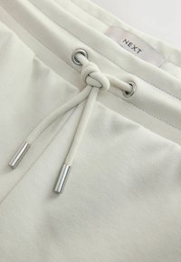 Gros plan sur la ceinture d’un pantalon de survêtement beige avec un cordon en tissu noué et des embouts métalliques argentés, affichant une étiquette de marque "NEXT" visible.