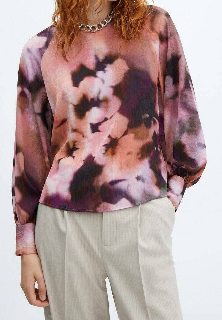 Personne portant un chemisier à manches longues tie-dye aux tons rose, violet et noir, associé à un pantalon beige clair et un collier chaîne épais.