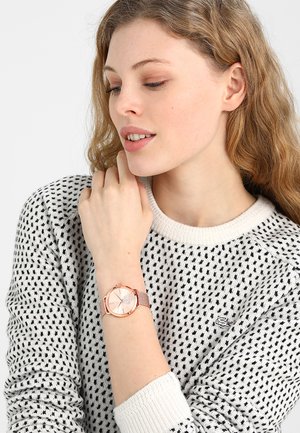 Femme aux cheveux châtain clair ondulés portant un pull blanc et noir à motifs, montrant une montre-bracelet en or rose à son poignet gauche.