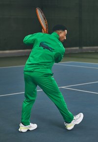 Zöld sportos tréningruha, textúrált felülettel, hátulján nagy kék krokodil logóval, fehér és sárga sportcipőkkel párosítva.