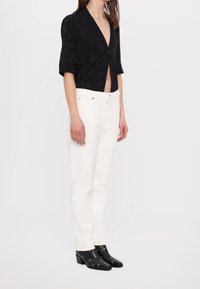 Schwarze, kurzes Bluse mit kurzen Ärmeln und V-Ausschnitt; kombiniert mit weißen, geraden Jeans. Schwarze Knöchelschuhe mit silbernen Ösenakzenten.