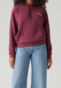 Persona vistiendo una sudadera maroon de Levi's con bolsillo frontal y jeans de pierna ancha de color azul claro, de pie frente a un fondo sencillo.