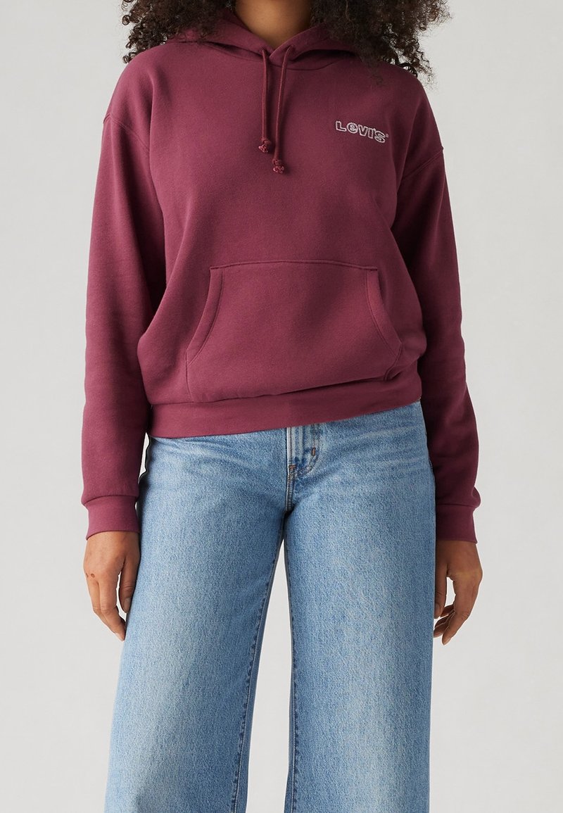 Persona vistiendo una sudadera maroon de Levi's con bolsillo frontal y jeans de pierna ancha de color azul claro, de pie frente a un fondo sencillo.