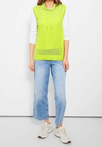 Felpa senza maniche verde con collo drappeggiato e lacci, indossata sopra una maglietta bianca a maniche lunghe. Abbinata a jeans larghi azzurri e sneakers.