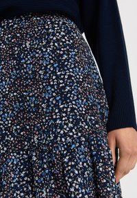 Spódnica w kwiatowy wzór w granatowym kolorze z małymi białymi, niebieskimi i różowymi kwiatami. Materiał jest tkaniną o miękkiej fakturze, z falbaniastym wykończeniem u dołu.