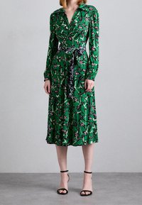 Zelené šaty s dlouhými rukávy, zavinovací design a květinový vzor v růžových a fialových akcentích. Detaily na pásku, délka pod kolena, kombinované s černými sandály.