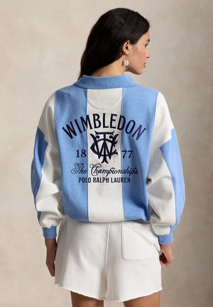 Femme aux cheveux foncés portant un sweat-shirt à rayures bleu et blanc avec le logo Wimbledon 1877 et un short blanc, vue de dos.