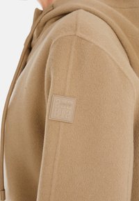 Helle braune Fleece-Hoodie mit strukturierter Oberfläche, verstellbarem Kordelzug und einem quadratischen Logopatch auf der linken Schulter.