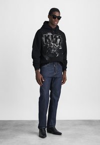 Čierna mikina s grafickou potlačou, modré džínsy a čierne kožené topánky. Model nosí čierne slnečné okuliare a stojí na neutrálnom pozadí.