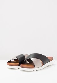 Schwarze und metallische Rutschen-Sandalen mit Kreuzriemen-Design, strukturierten silbernen Akzenten, braunem Fußbett und weißen, gerillten Sohlen.