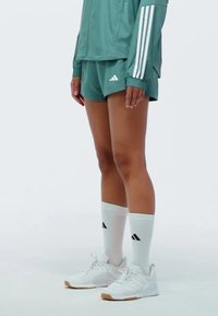 Sportliches Ensemble bestehend aus einer hellgrünen Jacke mit weißen Streifen, passenden Shorts und weißen Socken mit schwarzen Akzenten. Weiße Sneaker sind ebenfalls enthalten.