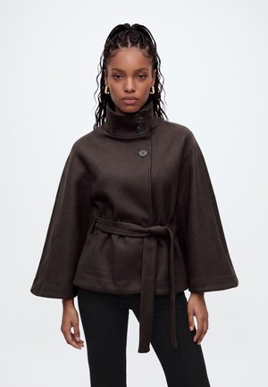 Femme aux cheveux tressés portant des boucles d'oreilles anneaux dorés et un manteau cape marron foncé ceinturé avec des boutons, posant devant un fond blanc.