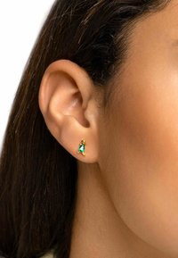 Orecchini dorati con una pietra verde a forma di goccia, caratterizzati da un design elegante, indossati sul lobo con la pelle liscia visibile.
