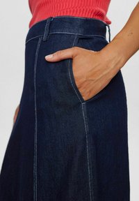 Tumši zilo džinsu svārku ar baltām dūrēm, ar sānu kabatu un cieši pieguļošu jostas vietu. Gluda tekstūra un A formas siluets.