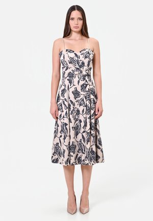 Femme portant une robe beige sans manches longueur genoux avec un imprimé floral noir et des talons hauts couleur nude, debout devant un fond blanc uni.