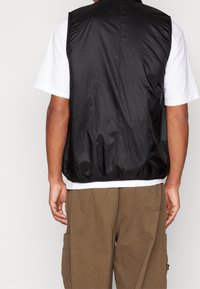 Gilet leggero nero con finitura testurizzata, orlo elastico e taglio dritto, indossato sopra una t-shirt bianca con pantaloni cargo marroni.