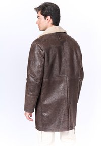 Abrigo de cuero marrón con un acabado texturizado, que presenta un collar de piel de oveja beige y un parche rectangular con logo en la parte posterior. Manga larga y corte recto.