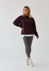 Bordowy sweatshirt z napisem "Księżniczka Pilates", nałożony na biały T-shirt, w połączeniu z szarymi legginsami i kremowymi sneakersami. Gładka faktura.