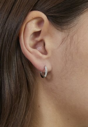 Kleiner silberner Creolen-Ohrring, verziert mit klaren Edelsteinen, an der linken Ohrmuschel einer Frau mit braunen Haaren.