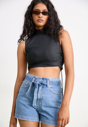 Zwarte mouwloze crop top met hoge hals en geplooide zijdetails, gecombineerd met lichtblauwe denimshorts met een gestrikt taille en opgerolde zoom.