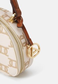 Kulatá kabelka z béžové texturované umělé kůže, zdobená zlatým vzorem s logem, s tmavě hnědou koženou rukojetí a akcentem se zlatým zipem.