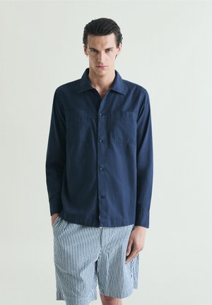 Hombre que lleva una camisa azul oscuro abotonada y pantalones cortos blancos con rayas verticales oscuras, de pie contra un fondo claro y liso.
