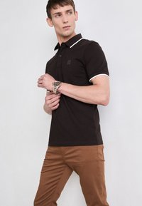 Fekete póló fehér gallér akcentusokkal, rövid ujjakkal és egy kis logóval a mellrészen, barna, slim fit nadrággal párosítva.
