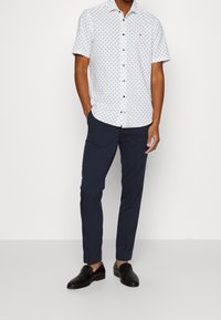 Camicetta bianca a maniche corte con motivo blu navy, chiusura a bottoni sul davanti, pantaloni blu scuro e scarpe slip-on in tessuto nero.