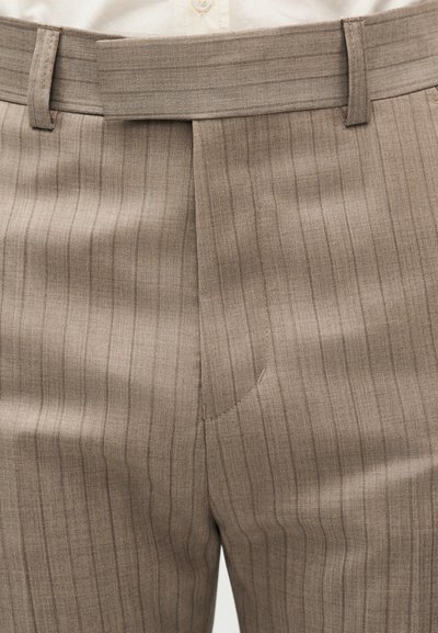 Μπεζ παντελόνι με ρίγες, ζωνάκι, θηλιές για ζώνη και λεπτομέρεια πιέτας μπροστά, φορεμένο πάνω από ένα λευκό πουκάμισο με κουμπιά.