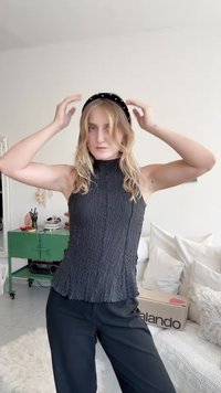 Černý texturovaný bezrukávový top s vysokým límcem a volánkovým lemem. Model nosí černou čelenku s bílými akcenty. Pozadí tvoří polštáře a zelená skříň.