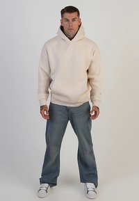 Felpa beige con cappuccio e tasca a marsupio, abbinata a jeans azzurri a gamba larga e sneaker bianche. Design minimalista, texture morbida.