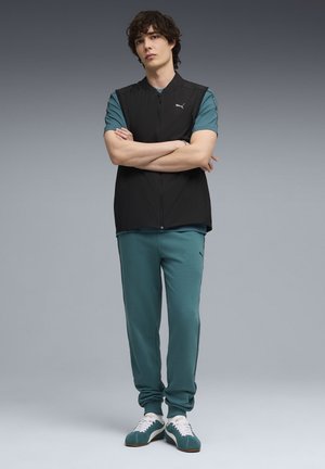 Zwarte mouwloze jas over een teal T-shirt met korte mouwen, gecombineerd met teal broek en groene sneakers. De jas heeft een Puma-logo.