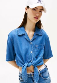 Camicetta di jeans blu a maniche corte con un davanti annodato, dotata di taschino sul petto e chiusura a bottoni, abbinata a jeans in denim a vita alta.