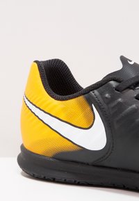 Zapatilla deportiva negra y amarilla con un acabado suave, que presenta un prominente logo en forma de swoosh blanco, patrones texturizados y un diseño de ajuste ceñido.