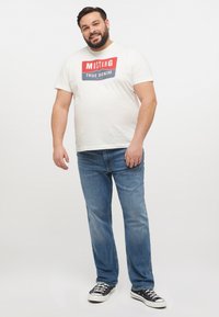 Vit T-shirt med röd och blå "MUSTANG TRUE DENIM" grafik, ihop med ljusblå jeans och svarta kanvas sneakers.