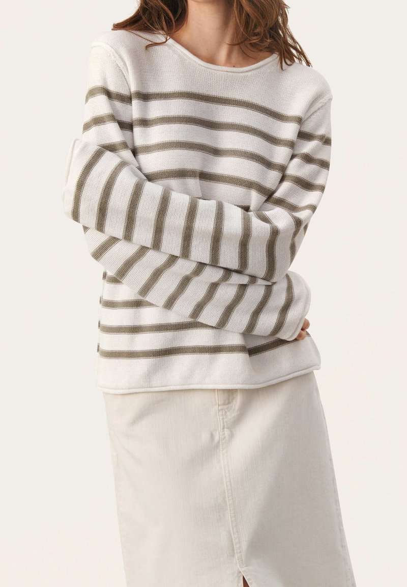 Pull rayé en beige clair avec des rayures vert olive, présentant une coupe ample, de larges poignets et un col rond, associé à une jupe légère.