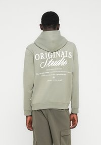 Άνδρας που φοράει ανοιχτό γκρι φούτερ με κουκούλα με το λευκό κείμενο «Originals Studio» στην πλάτη, συνδυασμένο με αντίστοιχο γκρι παντελόνι cargo, στέκεται μπροστά σε λευκό τοίχο.